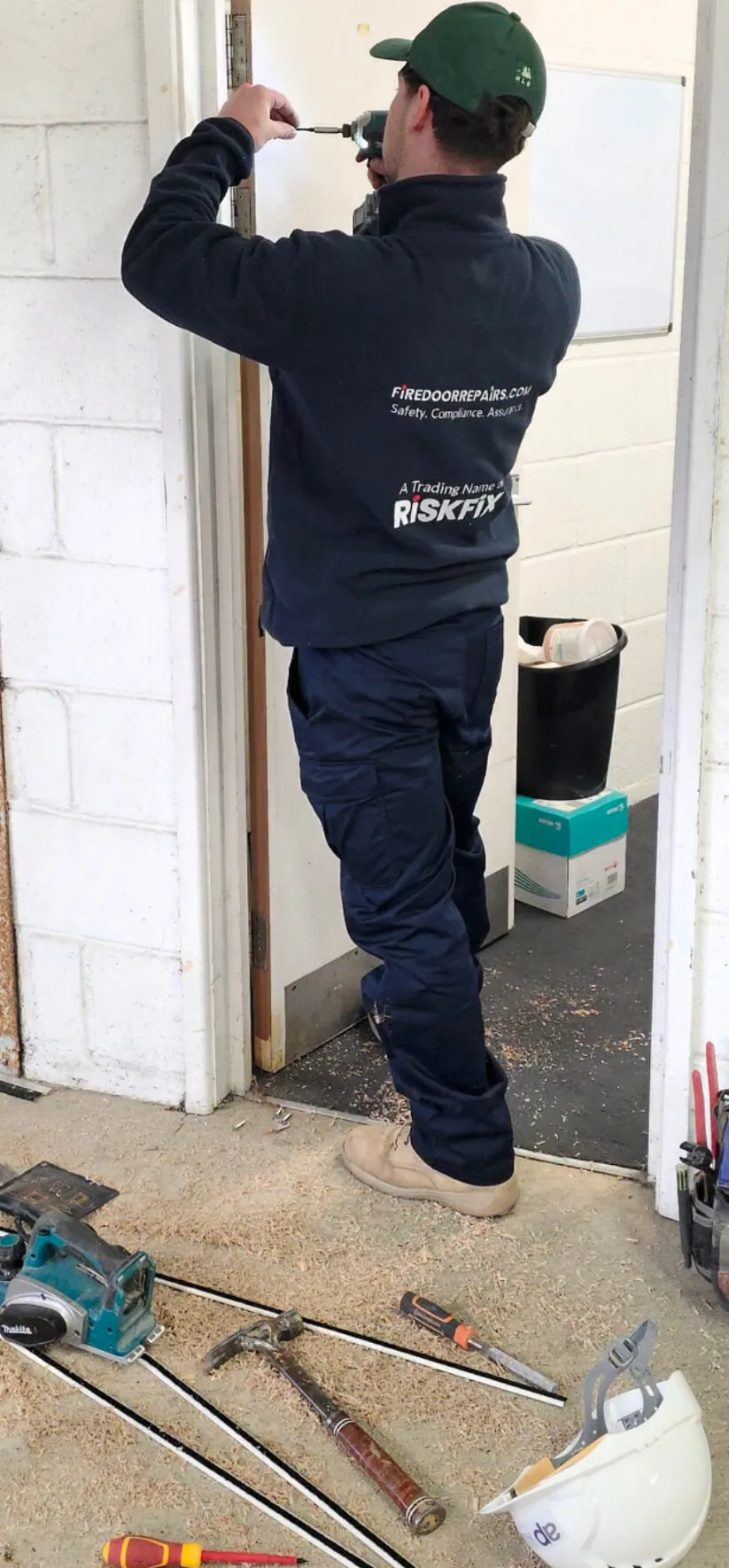 A worker using a cordless driver on the upper hinge of a fire door, with seals and tools laid out below