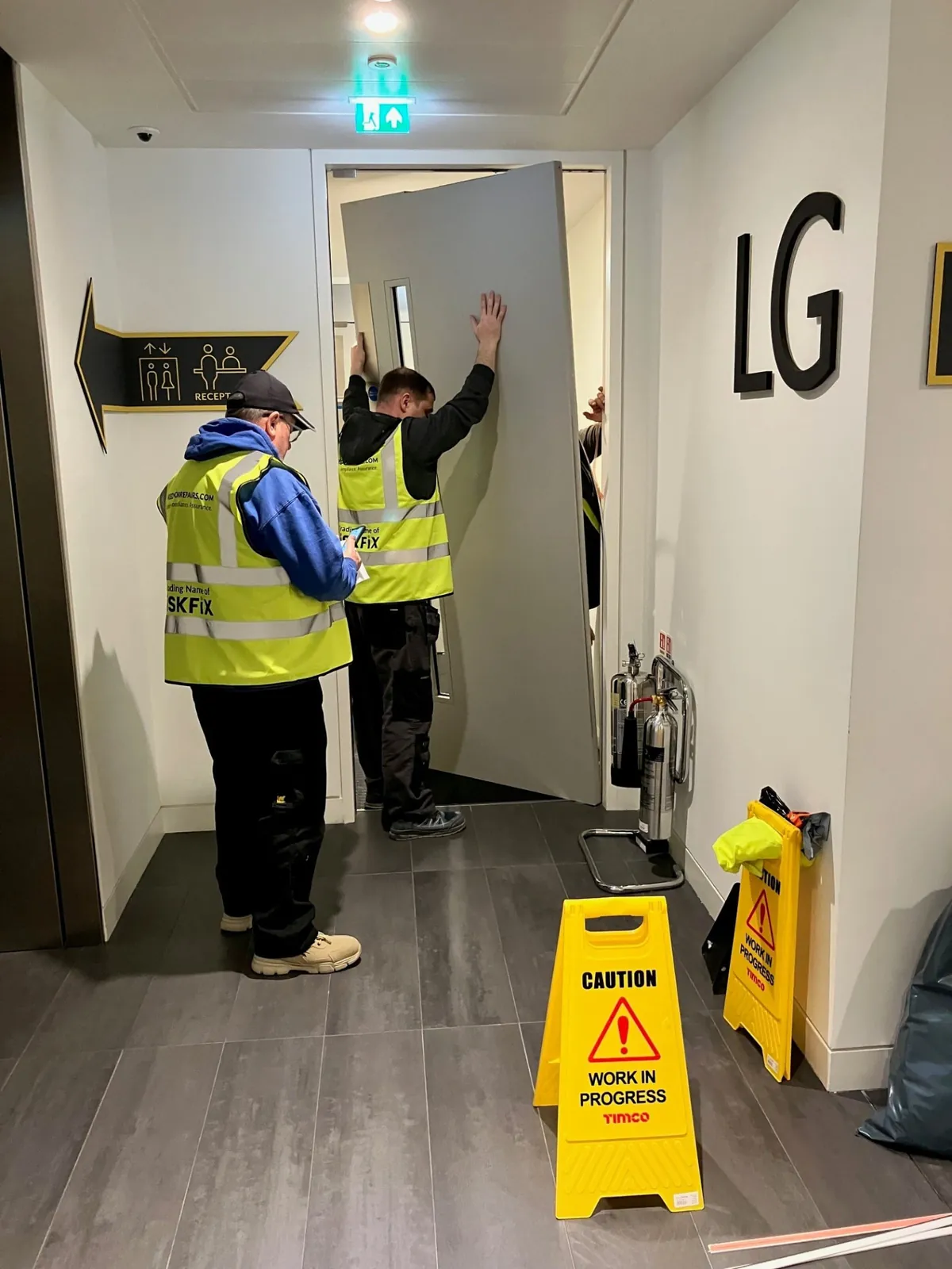 Two workers carrying a fire door leaf into a hallway where remedial works are in progress