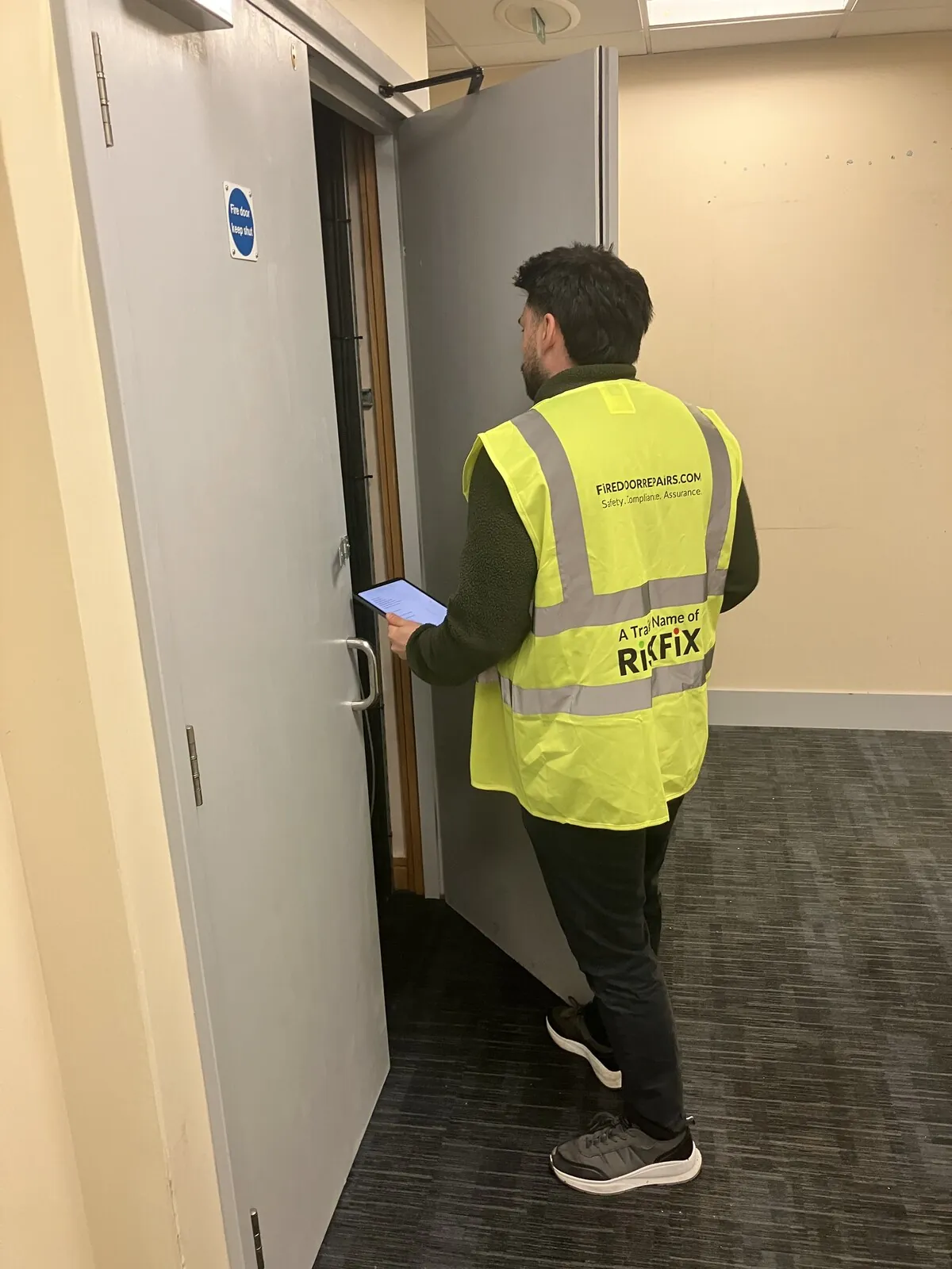 A surveyor holding a tablet while checking a pair of fire doors, one open to reveal a service riser